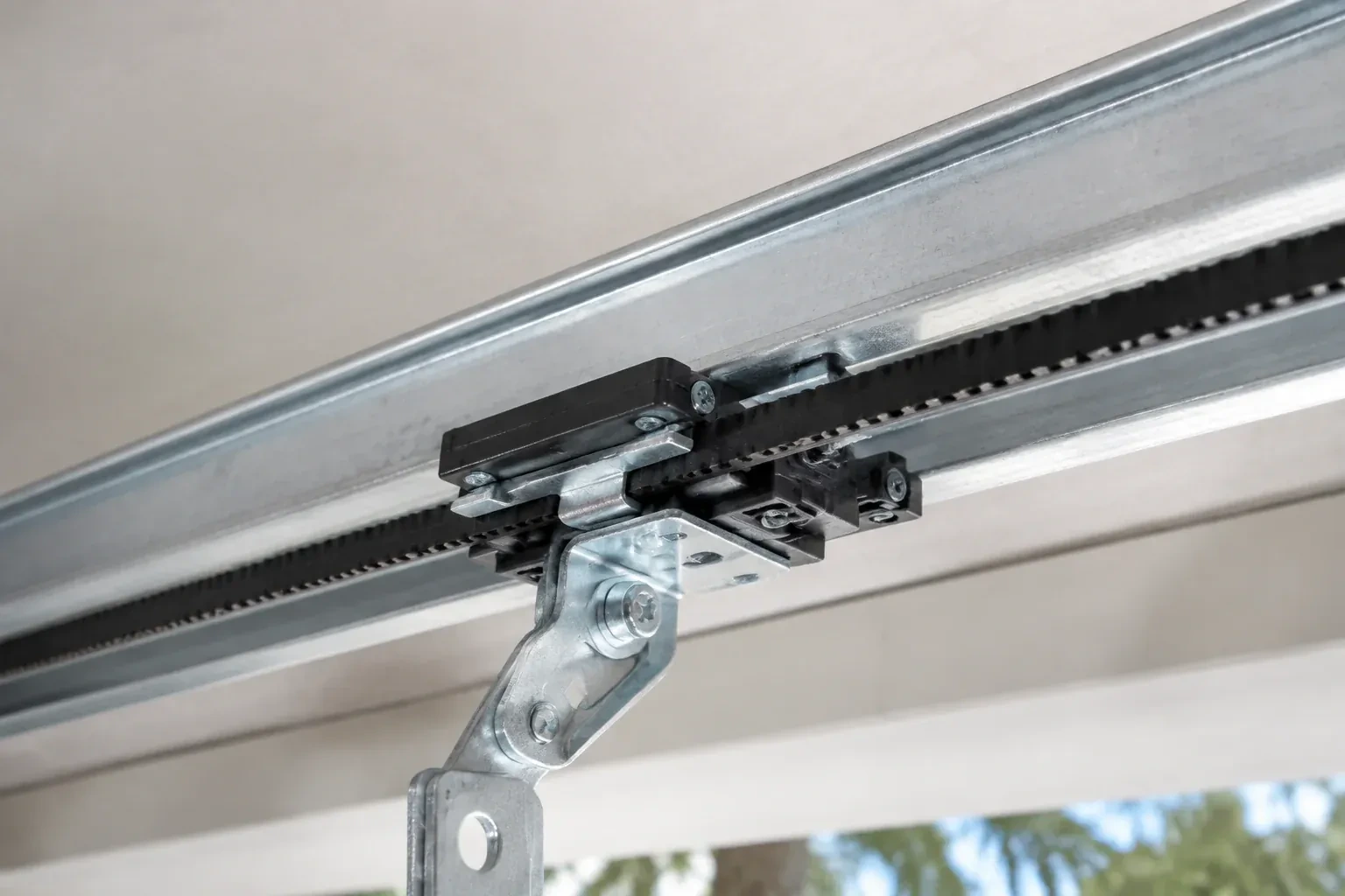Black plastic trolley and bent galvanized bracket on a steel opener rail with a toothed black belt, against a ceiling. — Belt-Drive Opener Repair, Richmond Hill