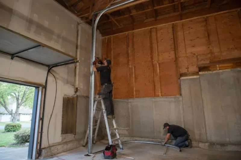 One person on a stepladder working beside a tall curved garage-door track while another kneels beside loose track sections in an unfinished garage. — High-Lift Garage Door Conversion, Richmond Hill
